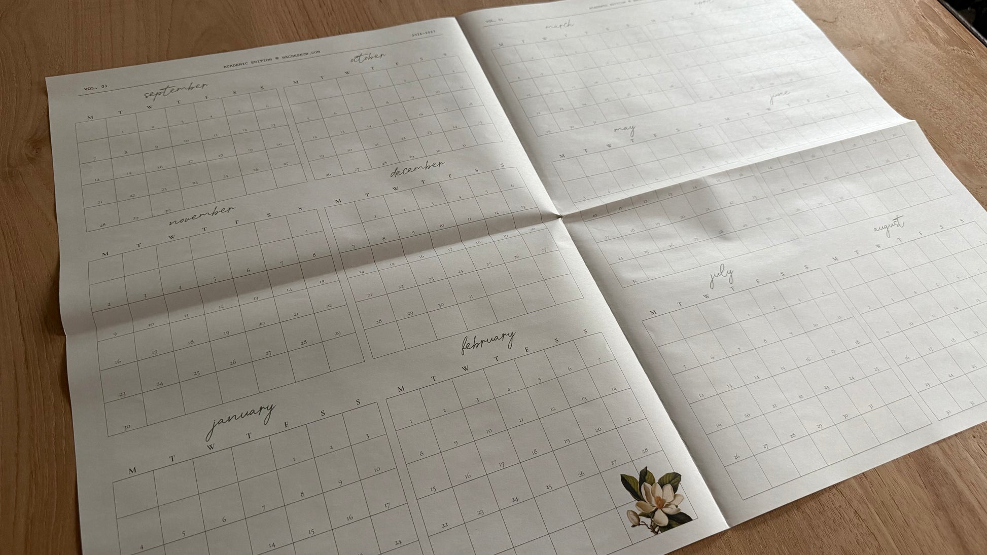 Fully opened Paper Calendar spread on a wood table showing all twelve months, September 2026 through August 2027, in Monday-start grids with a magnolia illustration in the corner.