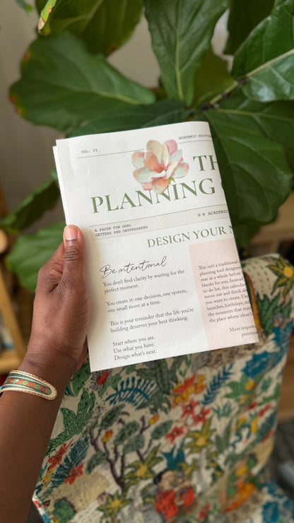 The Planning Calendar folded to page size and held in one hand, showing its scale against a fiddle leaf fig and patterned fabric.